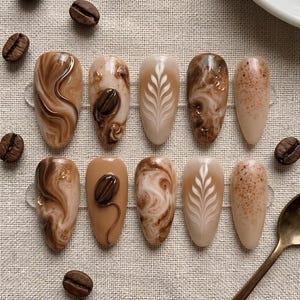 May include: A set of ten almond-shaped press-on nails featuring coffee-inspired designs. The nails display various patterns including brown and cream marble swirls, raised 3D coffee bean accents, white leaf latte art, and brown speckled textures.