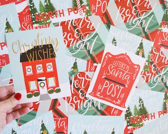 May include: A collection of Christmas cards featuring festive designs with red and green colors. The cards include illustrations of houses, mailboxes, and trees. The text on the cards includes "Greetings from the North Pole", "Merry & Bright", "Christmas Wishes", and "Letters to Santa Post".