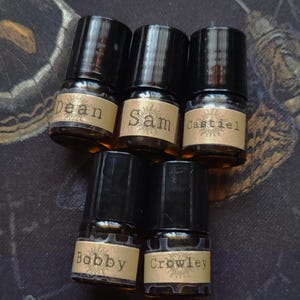 Peut inclure: Cinq petits flacons en verre ambré avec des bouchons noirs à vis. Chaque flacon présente une étiquette en papier beige avec une icône de soleil et les noms Dean, Sam, Castiel, Bobby et Crowley imprimés dans une police de style machine à écrire.