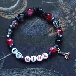Op de afbeelding: Een armband met rode, zwarte en heldere kralen. De armband spelt "BE MINE" met witte letterkralen. Een kleine bedel van een cocktailglas met een rode vloeistof hangt aan de armband.