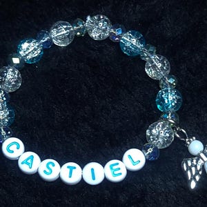 Könnte beinhalten: Ein blaues und klares Kristallperlenarmband mit einem silbernen Engelsanhänger und weißen Perlen, die den Namen "CASTIEL" buchstabieren.