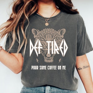 May include: A grey t-shirt featuring a graphic of a leopard's face and the text "DEF TIRED" in a metal band font. The text "POUR SOME COFFEE ON ME" is printed below the graphic.