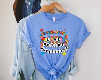 May include: A light blue short-sleeve t-shirt featuring a graphic print with the text LOVE ACCEPT UNDERSTAND in white circular bubbles. The design is surrounded by colorful puzzle pieces and floral shapes in red, yellow, and blue hues.