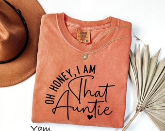 May include: A terracotta-colored short-sleeve t-shirt featuring the black printed text "OH HONEY, I AM That Auntie" with a small heart icon. The shirt has a crew neckline and a Comfort Colors brand label visible at the inner collar area.