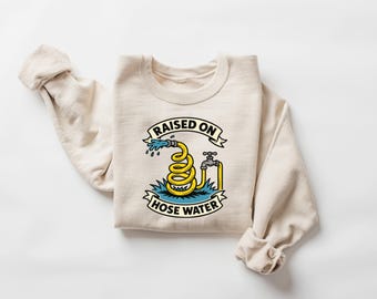 May include: A beige crewneck sweatshirt featuring a graphic illustration of a coiled yellow garden hose connected to a metal spigot, with blue water spraying out. The text "RAISED ON HOSE WATER" is printed in black capital letters on two banners.