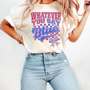 May include: A cream-colored t-shirt featuring a graphic print with the text "WHATEVER YOU SAY BLUE" in red and blue block letters. The design includes a red and blue checkered pattern, a blue script font, and a red and blue checkered bow graphic.