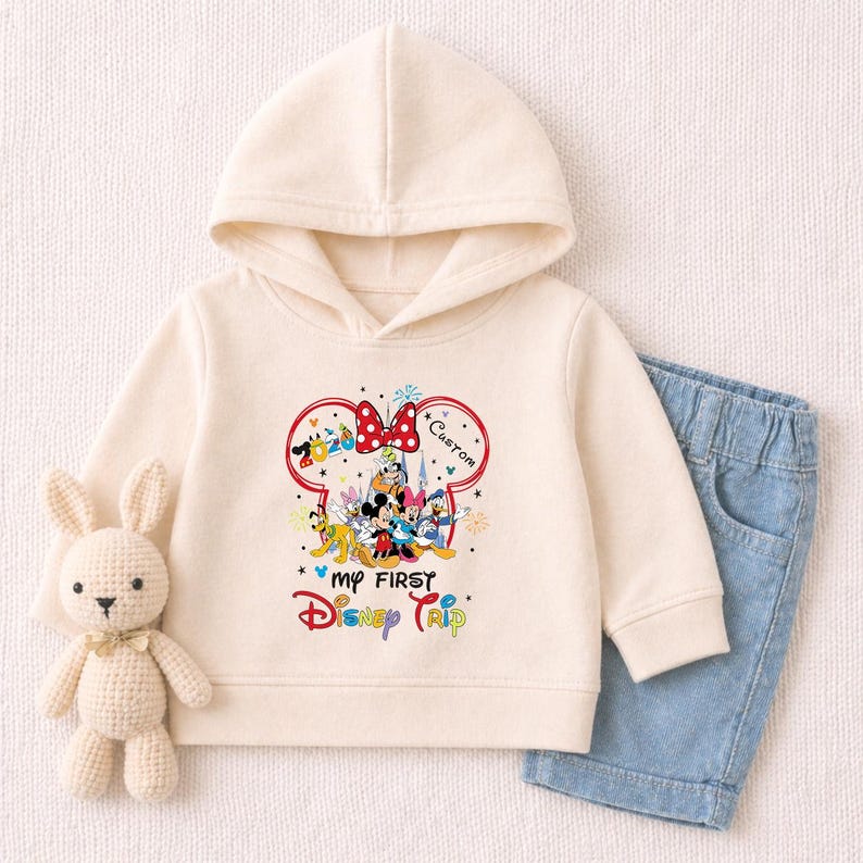 A white hoodie with a Disney character design and the text "My first Disney trip" printed on it, along with a pair of blue denim shorts and a crocheted bunny toy.