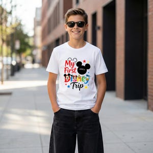 A young boy wearing a white t-shirt with a Disney-themed graphic and text, smiling and standing on a city sidewalk.