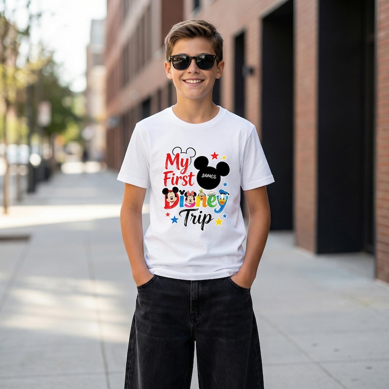 A young boy wearing a white t-shirt with a Disney-themed graphic and text, smiling and standing on a city sidewalk.