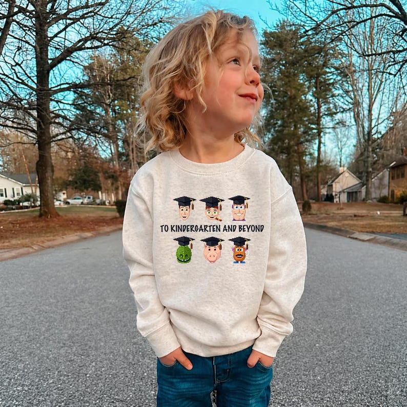 A young child wearing a white sweatshirt with a graphic design of cartoon faces and text, standing on a street with trees and houses in the background.
