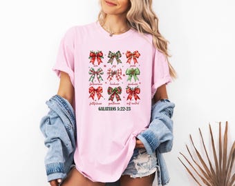 May include: A light pink t-shirt featuring a graphic of nine decorative bows in red, green, and plaid patterns. Each bow is labelled with a word such as love, joy, peace, and patience, with the text GALATIANS 5:22-23 printed at the bottom.