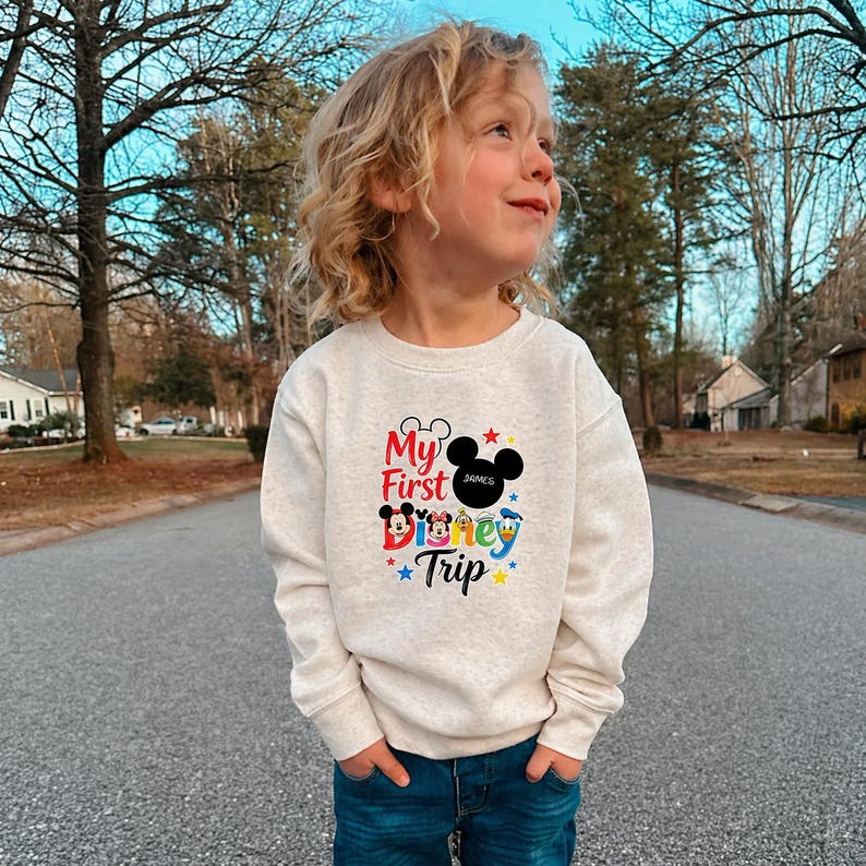 A young child wearing a white sweatshirt with a Disney-themed design stands on a street surrounded by trees.