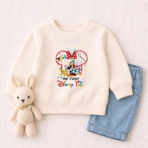 A white sweater with a Disney-themed design, a crocheted bunny toy, and a pair of blue denim shorts are arranged on a white background.