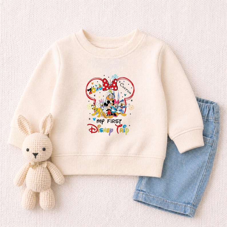 A white sweater with a Disney-themed design, a crocheted bunny toy, and a pair of blue denim shorts are arranged on a white background.