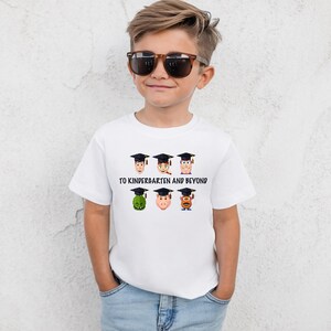A young boy wearing a white t-shirt with a graphic design of cartoon faces wearing graduation caps and the text "TO KINDERGARTEN AND BEYOND".