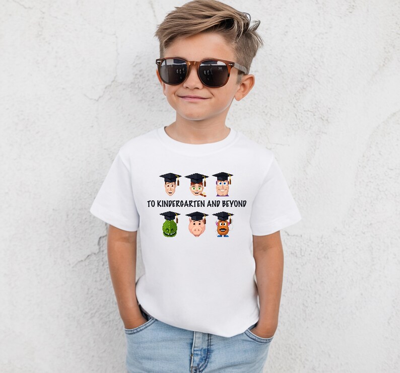 A young boy wearing a white t-shirt with a graphic design of cartoon faces wearing graduation caps and the text "TO KINDERGARTEN AND BEYOND".