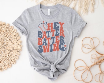 May include: A heather gray short-sleeve t-shirt featuring a graphic print with the text HEY BATTER BATTER SWING in wavy, stacked blue and red lettering. The design includes a baseball illustration and small star accents in blue and red.