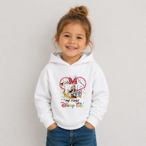 A young girl wearing a white hoodie with a Disney-themed design and text, smiling at the camera.