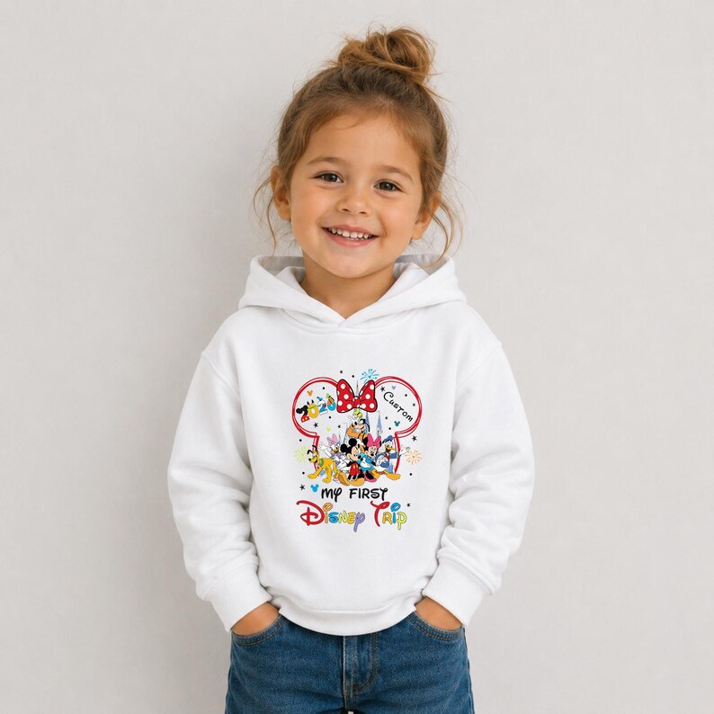 A young girl wearing a white hoodie with a Disney-themed design and text, smiling at the camera.