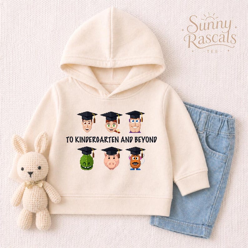 A white hoodie with a graphic design of cartoon characters wearing graduation caps, along with the text "To Kindergarten and Beyond". The hoodie is placed next to a pair of blue jeans, and a small stuffed rabbit is also present in the image.