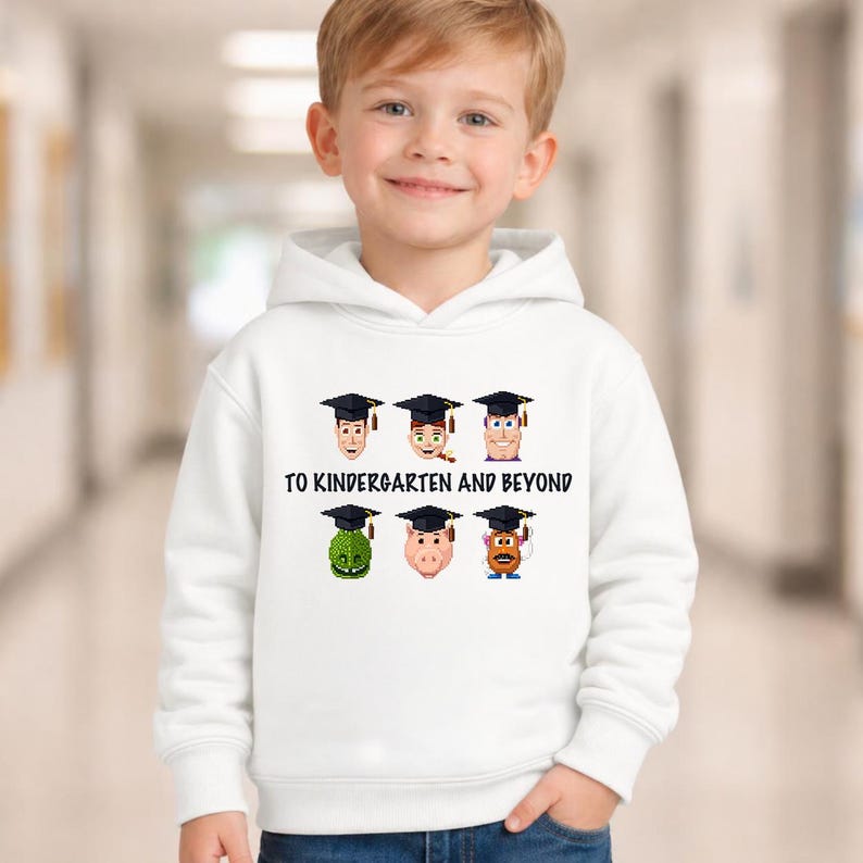 A young boy wearing a white hoodie with a graphic design of cartoon faces and text that says "TO KINDERGARTEN AND BEYOND".