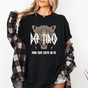 May include: Black t-shirt featuring a graphic of a leopard's face and the text "De-Tired" in a rock band style font. The text "Pour some coffee on me" is printed below the graphic.