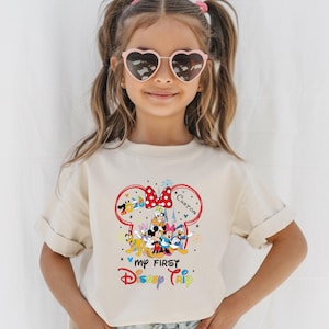 A young girl wearing a white t-shirt with a Disney-themed graphic and heart-shaped sunglasses, smiling at the camera.