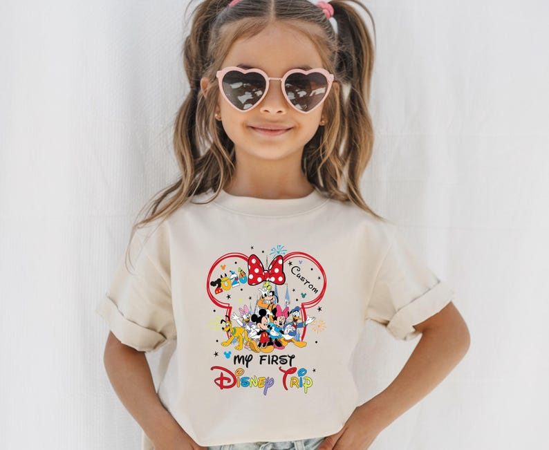 A young girl wearing a white t-shirt with a Disney-themed graphic and heart-shaped sunglasses, smiling at the camera.
