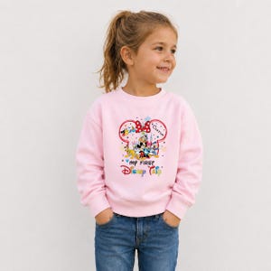 A young girl wearing a pink sweatshirt with a Disney character design and text on it, smiling and looking to the side.