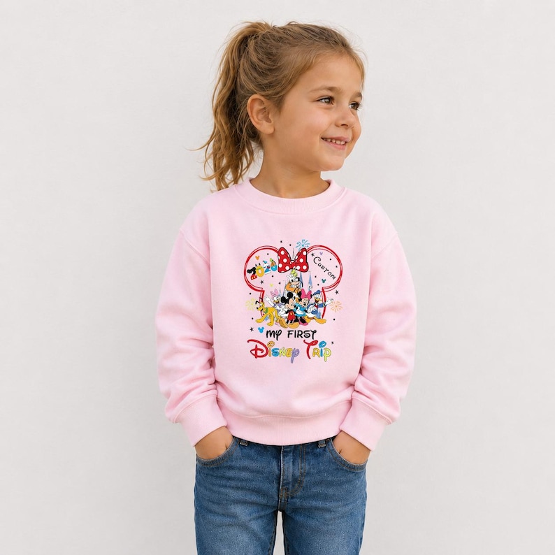 A young girl wearing a pink sweatshirt with a Disney character design and text on it, smiling and looking to the side.