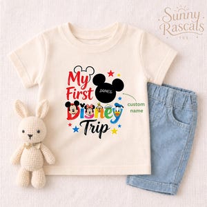 A white t-shirt with a Disney-themed graphic and text, along with a pair of blue denim shorts, and a stuffed bunny rabbit.