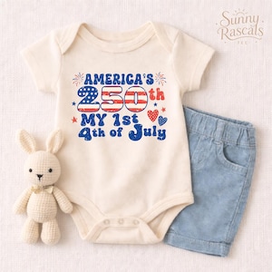 May include: A cream-colored short-sleeve baby bodysuit featuring the text America's 250th My 1st 4th of July in red and blue with a distressed American flag pattern on the numbers, accompanied by a small crocheted rabbit and light blue denim shorts.