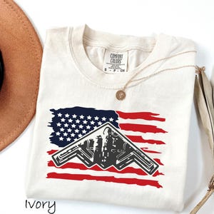May include: An ivory short-sleeve t-shirt featuring a graphic of a black B-2 stealth bomber silhouette layered over a distressed red, white, and blue American flag design. The shirt has a Comfort Colors brand label at the inner neckline.