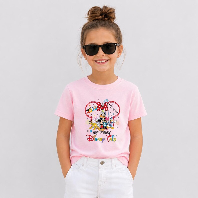 A young girl wearing a pink t-shirt with a Disney character design and sunglasses, smiling at the camera.