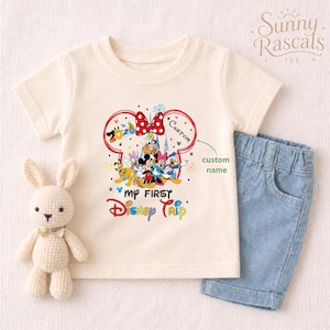 A white t-shirt with a Disney-themed graphic and a small stuffed bunny rabbit, along with a pair of blue denim shorts, are displayed on a white background.