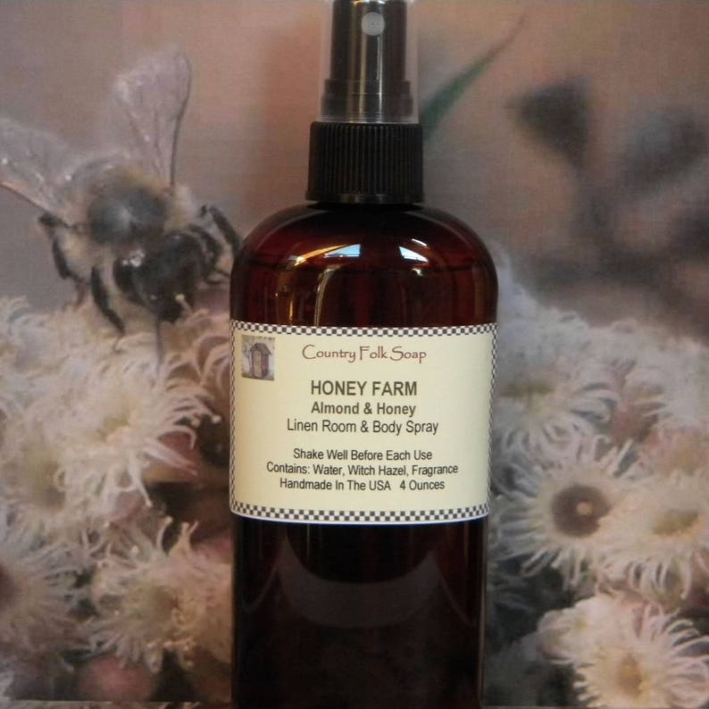 May include: A brown glass bottle with a black spray top. The bottle is labeled "Country Folk Soap Honey Farm Almond & Honey Linen Room & Body Spray". The label also includes the text "Shake Well Before Each Use Contains: Water, Witch Hazel, Fragrance Handmade In The USA 4 Ounces".