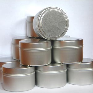 May include: A stack of round, silver-colored metal tins with flat lids. The containers feature a smooth, brushed finish on the top surfaces and a plain, cylindrical body design. These metallic storage vessels are arranged in a pyramid formation.