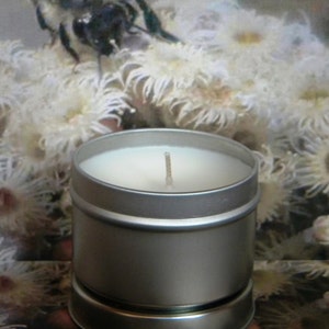 May include: A silver candle tin with a white candle and a single wick. The candle is surrounded by a white, fuzzy background.