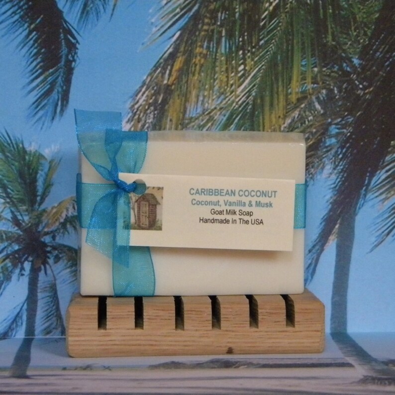 Pode incluir: Uma barra de sabonete de leite de cabra embrulhada em uma fita azul e uma etiqueta que diz "CARIBBEAN COCONUT Coconut, Vanilla & Musk Goat Milk Soap Handmade in The USA". O sabonete est&aacute; em um saboneteira de madeira.