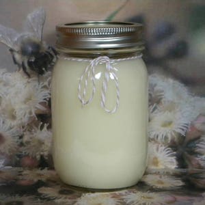 May include: A clear glass jar candle with a white lid and a twine bow. The candle is filled with a creamy white wax and is sitting on a floral background.