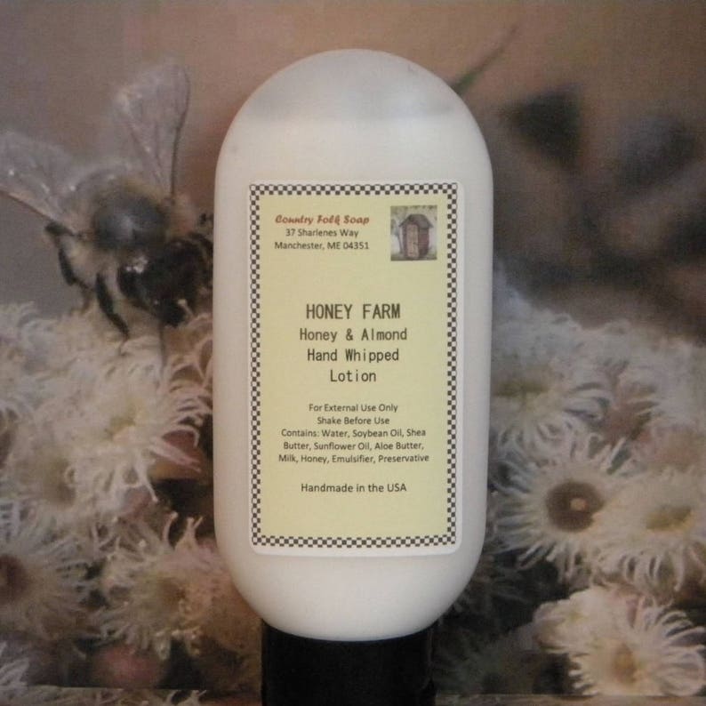 May include: A white bottle of Honey & Almond Hand Whipped Lotion by Honey Farm. The label is white with black text and a black and white checkered border. The bottle is sitting on a white surface with a bee and flowers in the background.