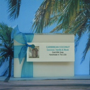 Pode incluir: Uma barra de sabonete branca embrulhada em uma fita azul com uma etiqueta que diz "CARIBBEAN COCONUT Coconut, Vanilla & Musk Goat Milk Soap Handmade in The USA".