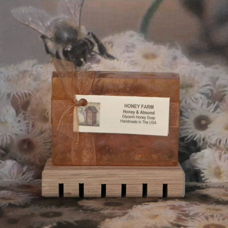 May include: A bar of Honey & Almond Glycerin Honey Soap from Honey Farm, handmade in the USA. The soap is a light brown color and is sitting on a wooden soap dish. The soap is wrapped in a gold ribbon and has a small tag attached to it.