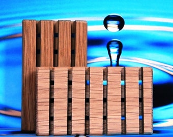 May include: Two wooden soap dishes with a slotted design, one rectangular and one square, are displayed against a blue background. The wood has a natural grain and is accented with dark lines. A water droplet is suspended above the dishes.