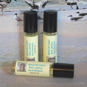 May include: Three small glass bottles with black roll-on tops and white labels. The labels have the text "BEACH BE CRAZY" and list the scent as "Sand, Jasmine, Sea & Mandarin Perfume Oil".
