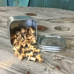 May include: A silver metal tin with a clear lid filled with wooden push pins. The tin is open and the push pins are spilling out onto a wooden surface.