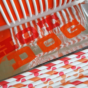 May include: A close-up of a silver foil package with the words "Fresh" and "Delicious" printed in orange on a white background. The package is partially covered by a red and white striped plastic container. The package is sitting on top of a stack of orange and white striped paper straws with red polka dots.
