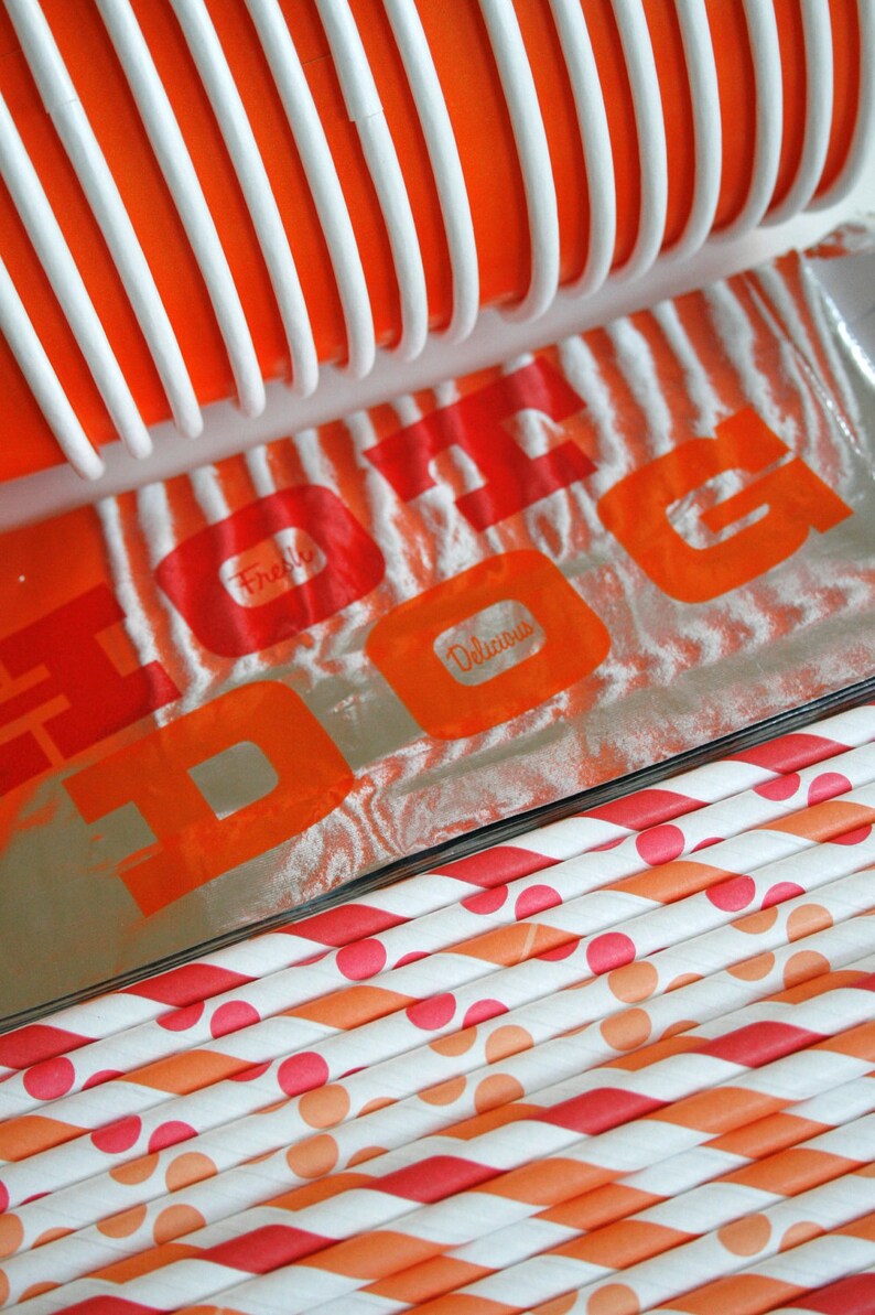 May include: A close-up of a silver foil package with the words "Fresh" and "Delicious" printed in orange on a white background. The package is partially covered by a red and white striped plastic container. The package is sitting on top of a stack of orange and white striped paper straws with red polka dots.