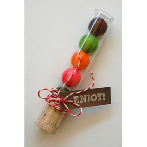 Pu&ograve; includere: Un tubo di vetro trasparente riempito con caramelle colorate, legato con uno spago rosso e bianco e un'etichetta marrone che dice "ENJOY!"