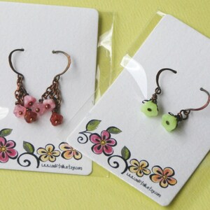 May include: Two pairs of earrings displayed on white cards with floral designs. One pair features pink flower-shaped beads and the other pair features green flower-shaped beads. Both pairs have copper-coloured hooks. The cards have the website www.selftakest.etsy.com printed on them.